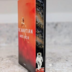 May include: A hardcover book titled The Martian by Andy Weir with a red dust jacket featuring an astronaut. The page edges are decorated with a black space scene, a galaxy of white stars, and a small white astronaut standing on a red surface.