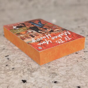 Puede incluir: Un libro de bolsillo titulado "If It Makes You Happy" con una cubierta y lomo naranja. La cubierta presenta una ilustraci&oacute;n colorida de personas y edificios. El logotipo de Penguin es visible. El libro est&aacute; sobre una superficie moteada.