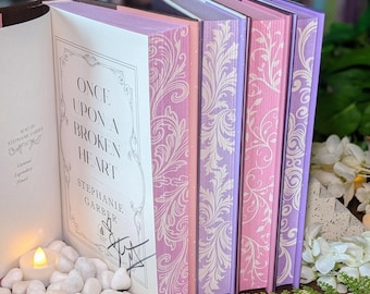 Puede incluir: Un conjunto de libros con bordes de página decorativos que presentan intrincados patrones florales blancos sobre fondos morados y rosas. El libro abierto muestra el título Once Upon a Broken Heart de Stephanie Garber con una firma en tinta negra.