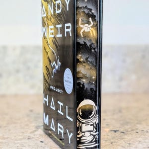 May include: A hardback book titled "Project Hail Mary" by Andy Weir. The book has a black cover with white and gold lettering and illustrations of an astronaut and space. The spine features an astronaut illustration.