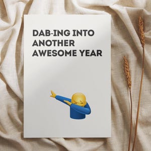 Puede incluir: Una tarjeta de felicitación blanca con el texto "DAB-ING INTO ANOTHER AWESOME YEAR" y un emoji amarillo haciendo el dab. El emoji lleva una camisa azul. La tarjeta está sobre un fondo de tela beige.