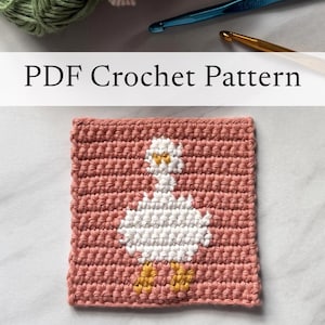 Peut inclure: Un carré au crochet représentant une oie blanche aux pattes jaunes sur un fond rose poudré. Le texte PDF Crochet Pattern est affiché au-dessus. Un crochet bleu de 7,0 mm et des pelotes de laine verte et rose sont visibles au-dessus.