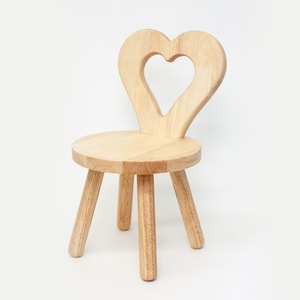 May include: A small, wooden chair with a heart-shaped backrest. The chair has a round seat and three legs. The wood has a natural, light brown color. The chair is designed for children.