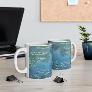 Puede incluir: Dos tazas de cer&aacute;mica blanca con un dise&ntilde;o de nen&uacute;fares azul y verde. Las tazas est&aacute;n sobre una superficie de madera clara.