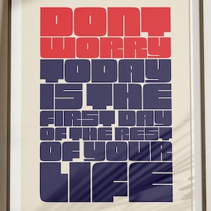 Puede incluir: Un póster motivacional con un marco negro y un fondo blanco. El texto dice "No te preocupes, hoy es el primer día del resto de tu vida." El texto está en una fuente en negrita, sans-serif, con las palabras "No te preocupes" en rojo y el resto en azul oscuro.