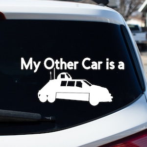 May include: A white car with a black rear window. The rear window has a white decal that says "My Other Car is a" and a silhouette of a car with antennas.
