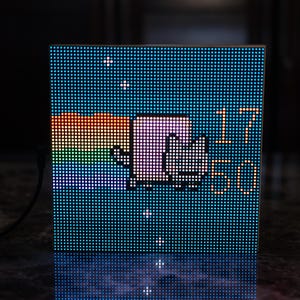 May include: A square LED display shows a pixelated image of a cat riding a rainbow, with the time 17:50 displayed in digital format. The background is a gradient of blue, with a black cable attached.
