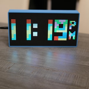 May include: A rectangular digital clock with a light blue frame and a black dot matrix display screen. The screen shows the time 11:19 PM in multicolored LED pixels, with a black power cord extending from the left side of the device.