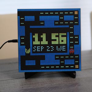 May include: A square digital clock with a pixelated blue maze background resembling a video game. The display shows the time 11:56 in lime green, with the date SEP 23 WE in white below. A black power cord extends from the left side of the device.