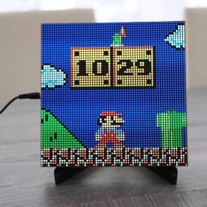 May include: A square LED pixel art display featuring a Super Mario theme with a blue background, green hills, and a character. The screen shows the time 10:29 inside two yellow blocks. The device is supported by a black stand and has a power cord.