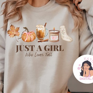 K&ouml;nnte beinhalten: Ein beiger Pullover mit einem grafischen Design aus herbstlichen Elementen, darunter ein K&uuml;rbis, Bl&auml;tter, eine Kerze, eine Tasse Kaffee und eine Socke. Der Text "Just a girl who loves fall" ist unter dem Grafikdruck angebracht. Ein kleiner Kreis mit dem Gesicht einer Frau und dem Text "Katco Digital Art" befindet sich in der unteren rechten Ecke.