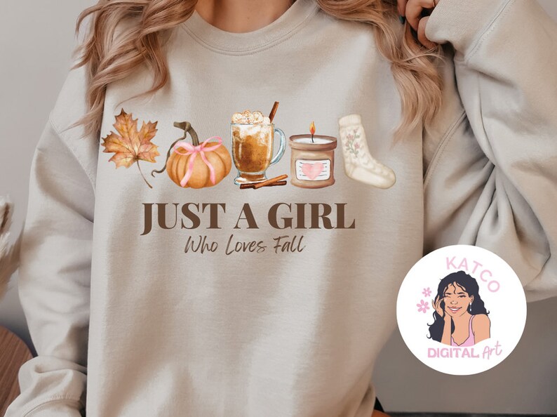 K&ouml;nnte beinhalten: Ein beiger Pullover mit einem grafischen Design aus herbstlichen Elementen, darunter ein K&uuml;rbis, Bl&auml;tter, eine Kerze, eine Tasse Kaffee und eine Socke. Der Text "Just a girl who loves fall" ist unter dem Grafikdruck angebracht. Ein kleiner Kreis mit dem Gesicht einer Frau und dem Text "Katco Digital Art" befindet sich in der unteren rechten Ecke.