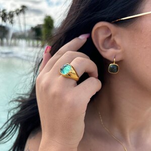 Puede incluir: Un anillo de oro con una gran piedra preciosa redonda de color verde. El anillo se lleva en el dedo índice de una mano. La persona también lleva un pendiente de oro con una piedra preciosa cuadrada de color verde oscuro.