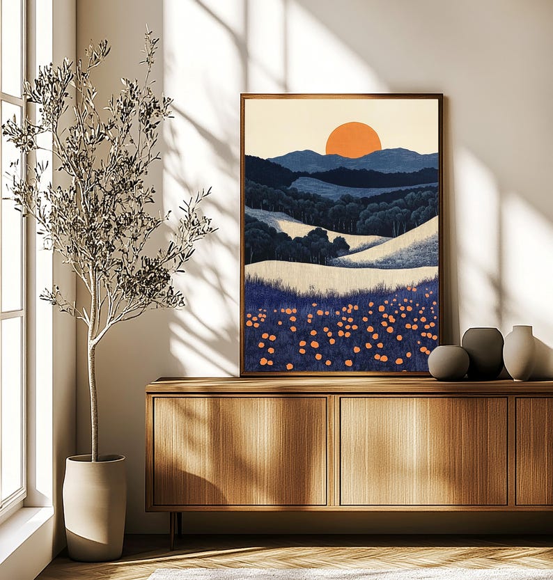 May include: A framed landscape art print featuring a stylized sunset over rolling hills and a field of orange flowers. The artwork has a warm color palette with shades of blue, orange, and cream. The frame is a warm brown color.