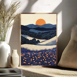 May include: A framed landscape art print featuring a stylized mountain scene with a large orange sun setting over blue hills and a field of orange flowers. The artwork is in a wooden frame and is displayed near a white vase with tall grass.