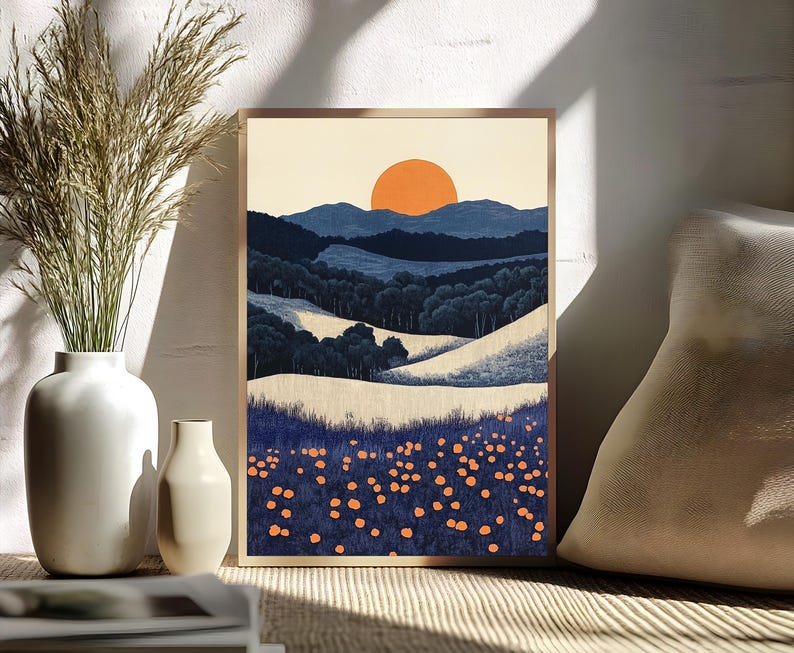 May include: A framed landscape art print featuring a stylized mountain scene with a large orange sun setting over blue hills and a field of orange flowers. The artwork is in a wooden frame and is displayed near a white vase with tall grass.