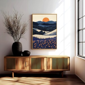 May include: A framed landscape art print featuring a stylized sunset over rolling hills and a field of orange flowers. The artwork is in a gold frame and hangs above a wooden cabinet with a vase of dried branches.