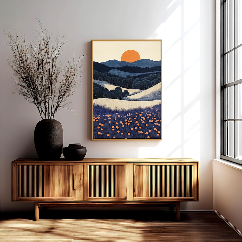 May include: A framed landscape art print featuring a stylized sunset over rolling hills and a field of orange flowers. The artwork is in a gold frame and hangs above a wooden cabinet with a vase of dried branches.