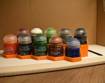 May include: A set of ten small paint bottles in a hexagonal orange plastic holder. The bottles are clear with various colored liquids inside. The labels on the bottles read "Citadel Colour" and list the paint color name.
