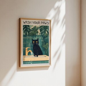 Puede incluir: Un gato negro con ojos amarillos se encuentra en un lavabo de ba&ntilde;o con un grifo dorado. El gato est&aacute; mirando a la c&aacute;mara. El lavabo es blanco con un salpicadero de azulejos verdes. El texto "WASH YOUR PAWS" est&aacute; escrito en letras negras sobre un fondo blanco en la parte superior de la imagen.