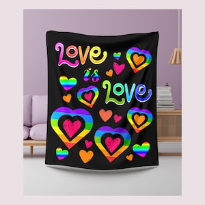 Könnte beinhalten: Schwarze Decke mit dem Schriftzug "Love is Love" in Regenbogenfarben, zusammen mit regenbogenfarbenen Herzen. Die Decke wird in einem Wohnzimmer gezeigt, über einem Sofa drapiert. Das Design ist lebendig und auffällig.