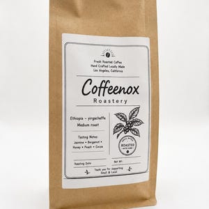 May include: A brown paper coffee bag with a white rectangular label. The label features the text Coeffenox Roastery, Ethiopia-yirgacheffe medium roast, and tasting notes of jasmine, bergamot, honey, peach, and cocoa, with a coffee plant illustration.