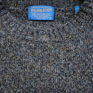 May include: Close-up of a gray and blue Pendleton sweater. The sweater has a crew neck and a blue label with white text that reads "PENDLETON 100% PURE VIRGIN WOOL PORTLAND, OREGON SINCE 1963". The sweater is made of a textured knit fabric.