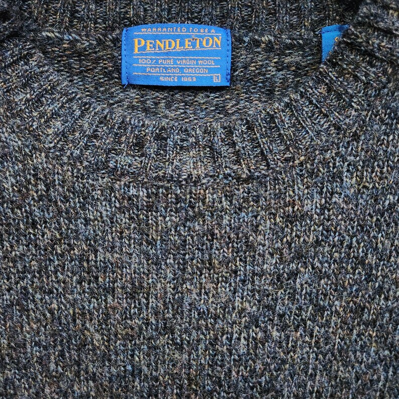 May include: Close-up of a gray and blue Pendleton sweater. The sweater has a crew neck and a blue label with white text that reads "PENDLETON 100% PURE VIRGIN WOOL PORTLAND, OREGON SINCE 1963". The sweater is made of a textured knit fabric.