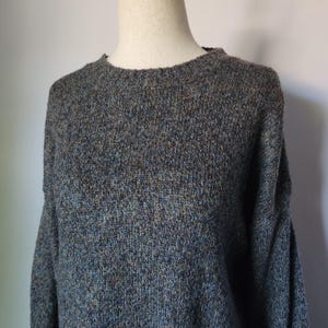 May include: A close-up of a gray, crew neck sweater. The sweater has a textured knit pattern with flecks of blue, brown, and tan throughout. The sleeves are long and the neckline is ribbed.