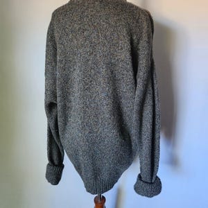 May include: A gray, textured sweater with long sleeves and ribbed cuffs. The sweater has a crew neck and a slightly oversized fit. The fabric appears to be a blend of gray, brown, and blue yarns, creating a speckled effect.