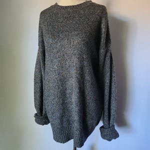 May include: A gray, oversized sweater with a crew neck and long sleeves. The sleeves have folded cuffs. The sweater is made of a textured knit fabric and is displayed on a mannequin.