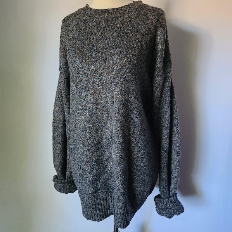 May include: A gray, oversized sweater with a crew neck and long sleeves. The sleeves have folded cuffs. The sweater is made of a textured knit fabric and is displayed on a mannequin.