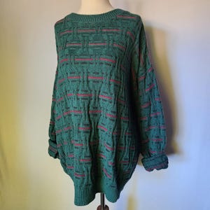 May include: A long-sleeve knit jumper in a deep forest green hue, featuring a prominent raised basket-weave texture throughout the body and sleeves, accented by thin, horizontal stripes of vibrant red yarn woven into the pattern.