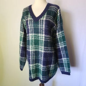 May include: A long-sleeve V-neck sweater featuring a plaid pattern in shades of dark green, navy blue, and white. The knit garment has a ribbed neckline, cuffs, and hem, with a consistent tartan-style check design throughout the entire piece.