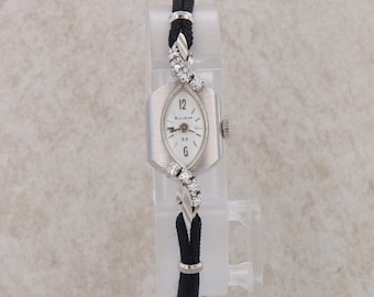 May include: A silver Bulova wristwatch with a white face and diamond accents on the band. The watch has a black band and a rectangular face with an oval-shaped window.