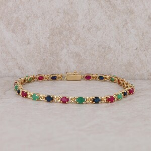 May include: A gold bracelet featuring a series of oval-cut gemstones in alternating colors of red, green, and blue. The bracelet is secured with a gold clasp and is displayed on a neutral background.