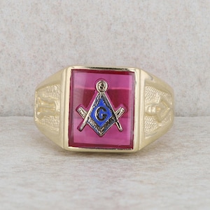 May include: A gold ring with a red stone featuring a blue and gold Masonic symbol.