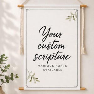 Könnte beinhalten: Ein weißer Wandbehang aus Stoff, der an einer Holzstange mit einem cremefarbenen Seil befestigt ist. Das Banner zeigt den schwarzen Text "Your custom scripture" und "VARIOUS FONTS AVAILABLE" mit grünen Aquarell-Blattillustrationen.