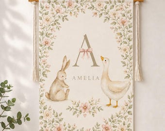 May include: A rectangular wall hanging featuring a watercolor illustration of a rabbit and a goose surrounded by a floral border. The center displays the letter A with a pink bow and the name AMELIA in capital letters. It hangs from wooden rods.