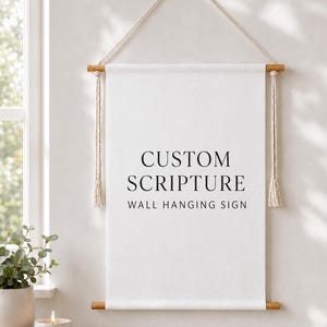 Puede incluir: Un cartel de tela blanca para colgar en la pared, suspendido de un listón de madera con un cordón beige. El cartel presenta el texto impreso en negro CUSTOM SCRIPTURE WALL HANGING SIGN. La tela se sujeta con listones de madera con borlas.