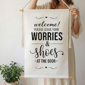 Könnte beinhalten: Ein cremefarbenes Wandbehang aus Baumwolle mit dem Text "welcome! PLEASE LEAVE YOUR WORRIES & SHOES AT THE DOOR." Das Schild hat schwarze Schrift und Holzdübel. Verfügbare Größen: 20x30 cm, 30x46 cm, 41x61 cm, 51x76 cm und 61x91 cm.