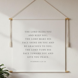Peut inclure: Une tenture murale avec le texte "THE LORD BLESS YOU AND KEEP YOU; THE LORD MAKE HIS FACE SHINE ON YOU AND BE GRACIOUS TO YOU; THE LORD TURN HIS FACE TOWARD YOU AND GIVE YOU PEACE. NUMBERS 6:24-26". En dessous, un meuble en bois avec une plante.