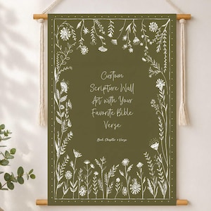以下が含まれることがあります： 白い花のイラストと「Custom Scripture Wall Art with Your Favorite Bible Verse」のテキストが特徴のオリーブグリーンのウォールアート。木製の棒と天然のロープで吊るされています。