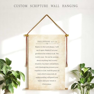 Puede incluir: Un colgante de pared con escritura personalizada con texto de Filipenses 4:7-13. El rollo beige está suspendido por un cordón de cuero marrón y clavijas de madera. La imagen incluye una mesa de madera con plantas en jarrones y macetas.