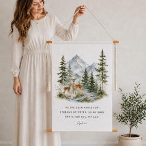 Könnte beinhalten: Ein vertikaler Wandbehang aus Stoff mit einer Aquarellillustration von zwei Hirschen in einem Wald mit Bergen im Hintergrund. Der Text lautet: AS THE DEER PANTS FOR STREAMS OF WATER, SO MY SOUL PANTS FOR YOU, MY GOD. Psalm 42:1. Holzstäbe.