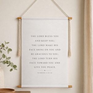 May include: A white wall hanging with wooden dowels and a rope hanger. The text reads "The Lord bless you and keep you; the Lord make his face shine on you and be gracious to you; the Lord turn his face toward you and give you peace." Numbers 6:24-26.