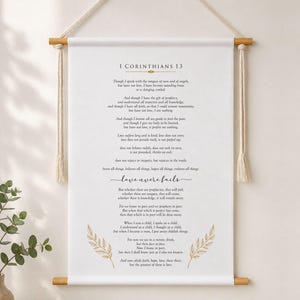 May include: A white fabric wall hanging featuring the text of 1 Corinthians 13 in black font with gold accents. The banner is suspended from a light-coloured wooden rod with a cream-coloured rope and includes decorative gold leaf illustrations.