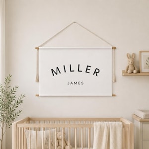 Puede incluir: Un estandarte de pared de tela blanca con el texto negro MILLER JAMES en una fuente sans-serif clara. El estandarte está suspendido de una varilla de madera de color claro con una cuerda a juego y borlas decorativas en cada lado.
