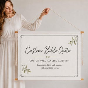 May include: A rectangular cotton wall hanging tapestry suspended from a wooden rod with a string. The off-white fabric features a black border, green leaf illustrations, and the text: Custom Bible Quote, COTTON WALL HANGING TAPESTRY.