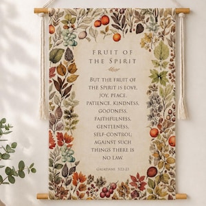 Puede incluir: Un tapiz rectangular de tela colgado de una varilla de madera con una cuerda color crema. El centro presenta el texto Fruit of the Spirit, Galatians 5:22-23, rodeado por un borde botánico de hojas de otoño coloridas, bayas y frutas.